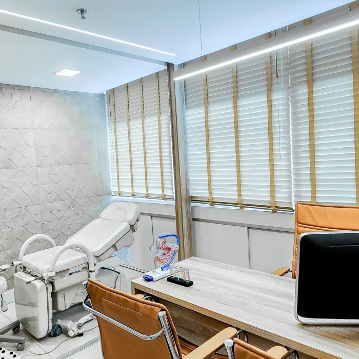 Sala de atendimento médico moderna e bem iluminada, com maca ginecológica, cadeira giratória, mesa de atendimento com computador, cadeiras para pacientes e persianas de madeira cobrindo janelas amplas.