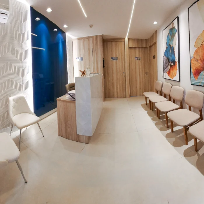 Recepção de clínica médica elegante com poltronas estofadas alinhadas, balcão de atendimento em madeira clara, ar-condicionado, paredes com acabamento texturizado e quadros decorativos coloridos na parede.